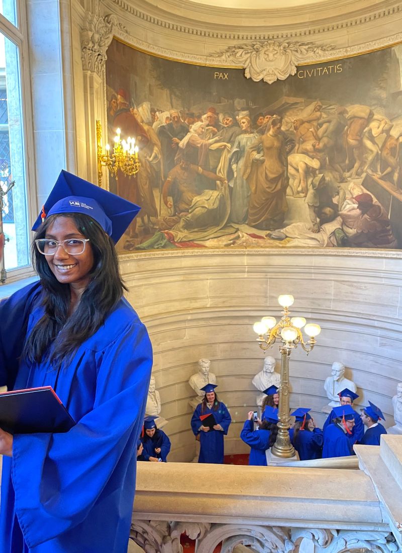 Nadia Ramdhanie in a blue VUB graduation cap and gown, smiling, in a grand muraled university hall.